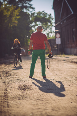 Sylwetka Andrzeja Pągowskie sfotografowana od tyłu w pozie kowboja z aparatem w dłoni w tle pani na rowerze